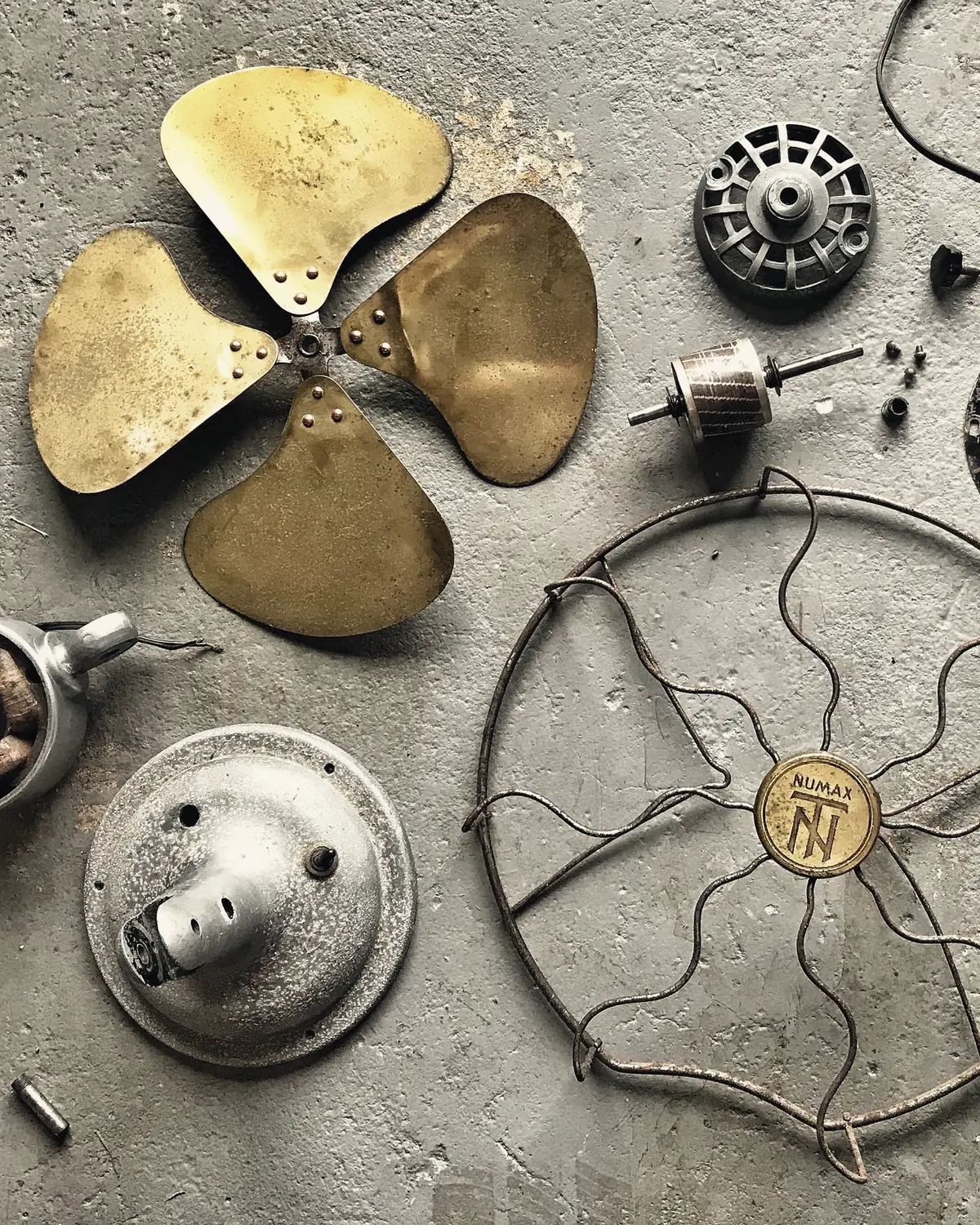 The parts of a vintage electric fan laid out on a concrete floor: blades, motor, cage and screws