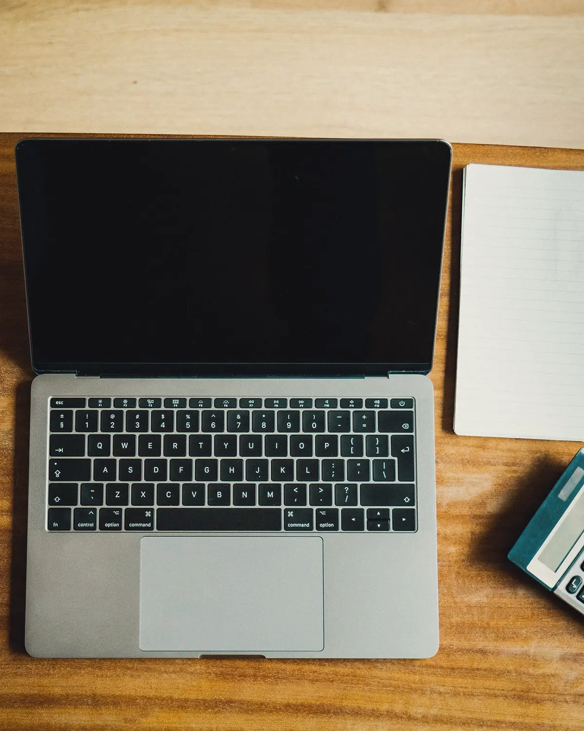 A laptop, an open notebook and a calculator on a wooden desk, from above